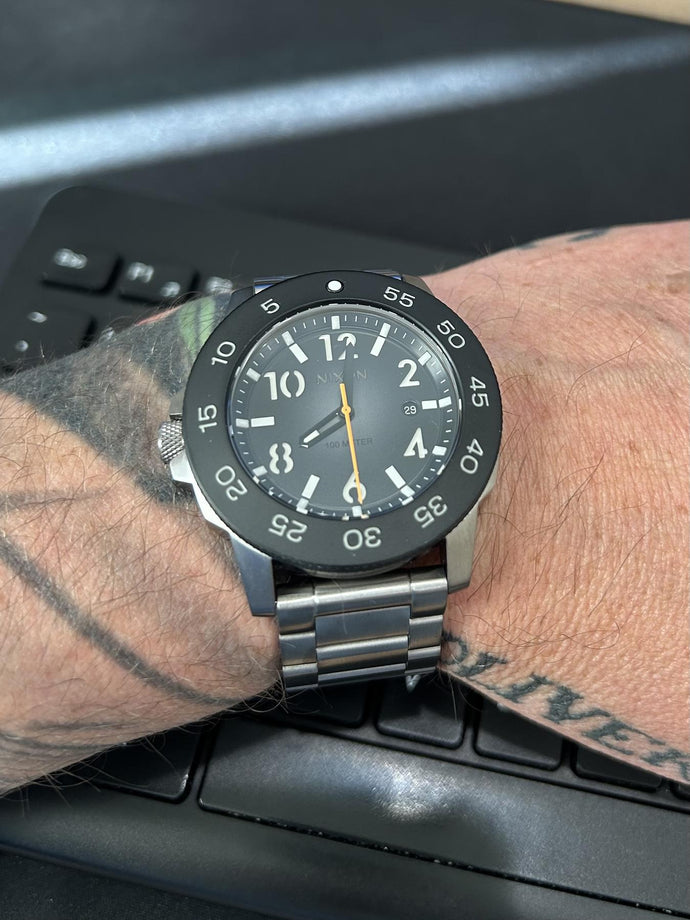 Black wristwatch on a person's wrist with tattoos, wearing a black shirt.