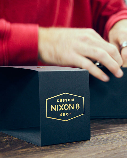 Nixon branded box on a wooden surface with a person wearing a red shirt in the background.