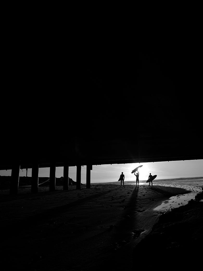 Silhouettes of people on a beach at sunset or sunrise, with a pier in the foreground.